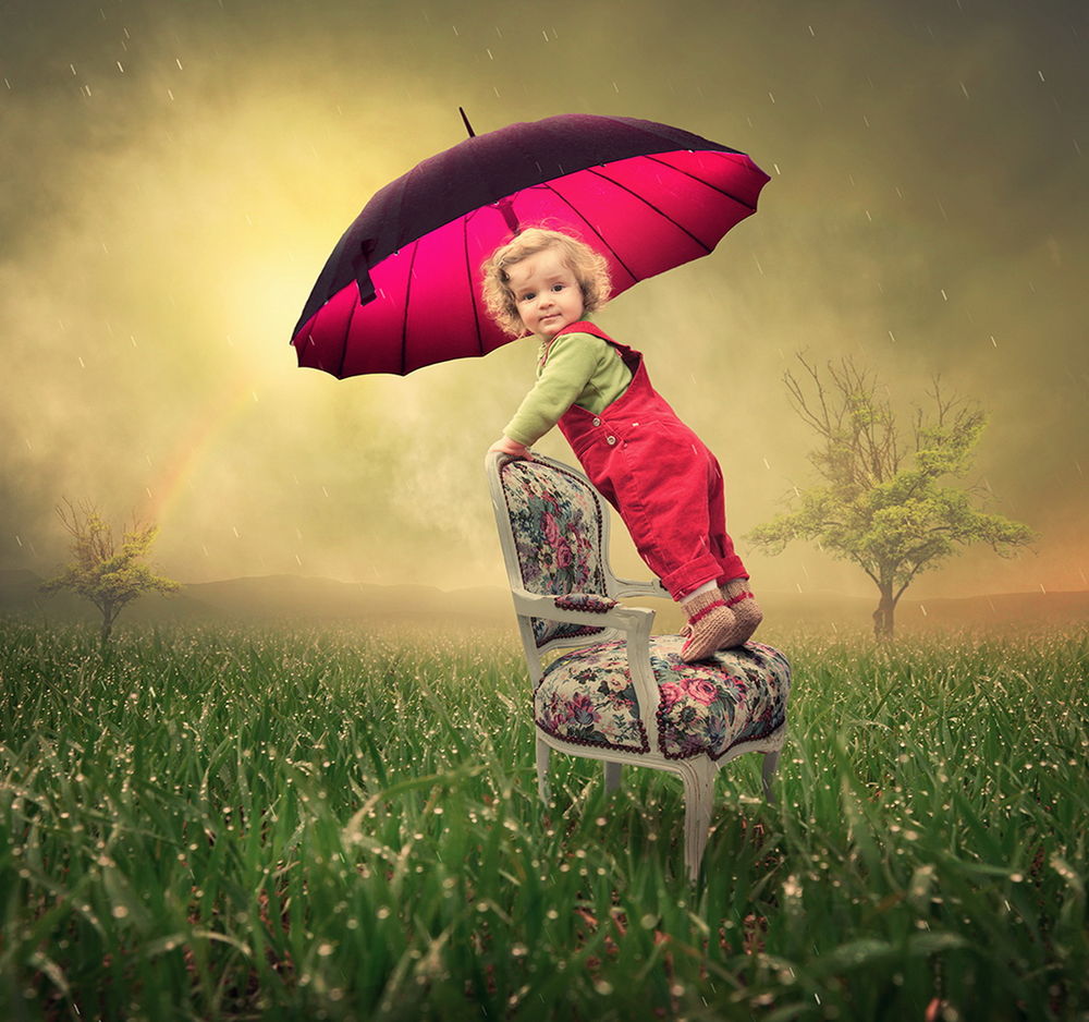 Обои для рабочего стола Белоголовый кудрявый ребенок, одетый в красный комбинезон, стоящий на мягком стуле с цветной обивкой среди зеленой травы, держит в руках красный зонтик, закрываясь им от сильного дождя на фоне пасмурного неба в туманной дымке и появившейся радуги, фотография Garas lonut