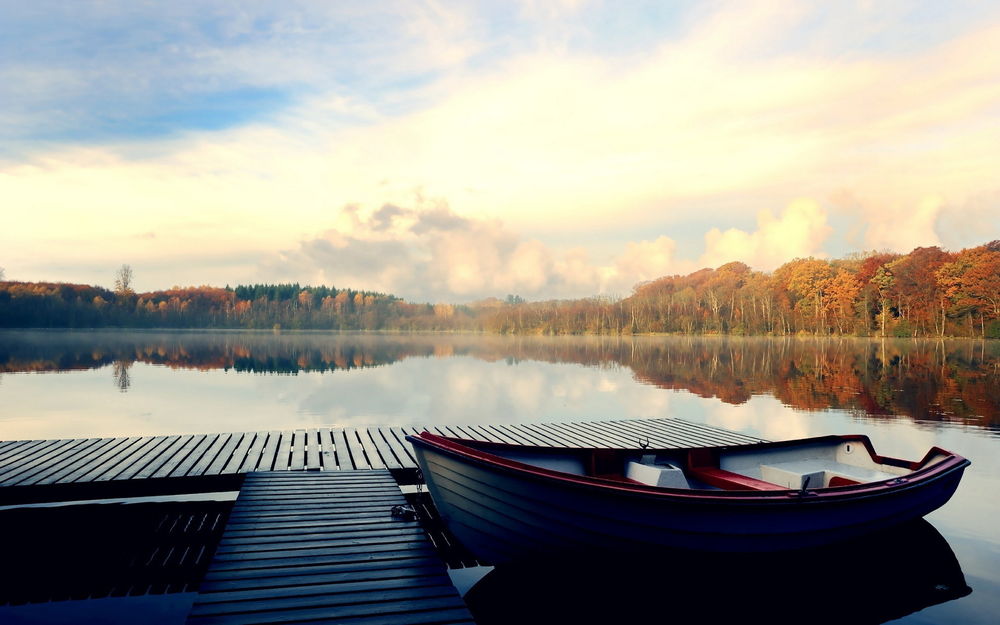 Обои для рабочего стола Лодка пришвартованная у небольшого деревянного мостика в озере, по берегам растут осенние деревья и легкий туман стелется по водной глади