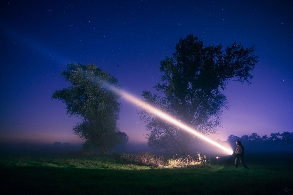 Обои для рабочего стола Мужчина, стоящий на поляне возле дерева, направил яркий луч света от электрического фонаря в сторону ночного звездного неба, фотография Wojeiech Grzanka