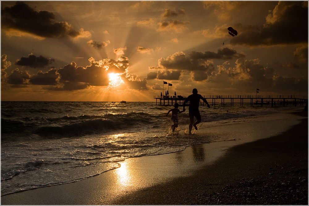 Обои для рабочего стола Отец с сыном, бегущие по песчаному пляжу морского берега на фоне золотистого заката солнца, парящего в небе парашютиста, фотография Aleetet