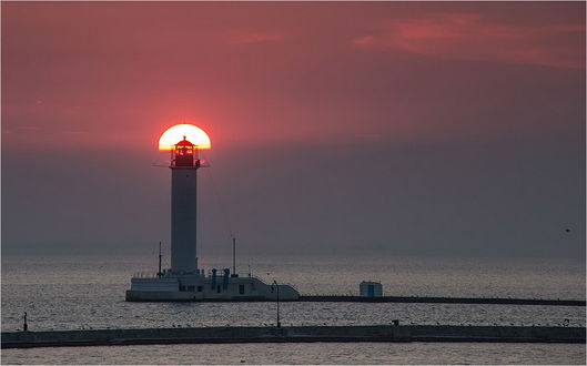 Обои на рабочий стол Маяк, стоящий в прибрежных морских водах на фоне заходящего за темные тучи ярко светящегося солнца, туманной линии горизонта, фотография Сергея Дымченко