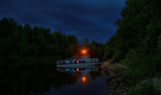 Обои на рабочий стол Грузовая баржа, стоящая у каменистого берега реки, с растущими на нем кустарниками и деревьями, над палубой горит ярко светящийся красный фонарь, отражающийся в воде на фоне пасмурного ночного неба, фотография optimist