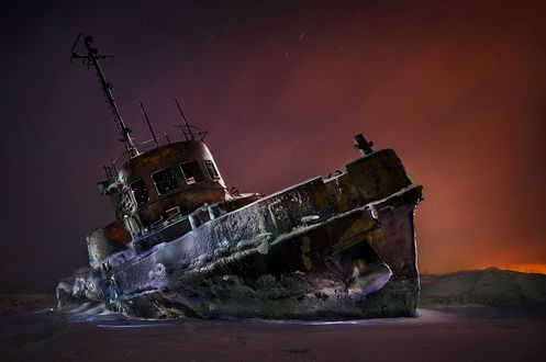 Обои на рабочий стол Ветхий, заброшенный рыболовный баркас, вмерзший в лед возле речного берега на фоне вечернего зимнего заката, фотография Руслана Бабина