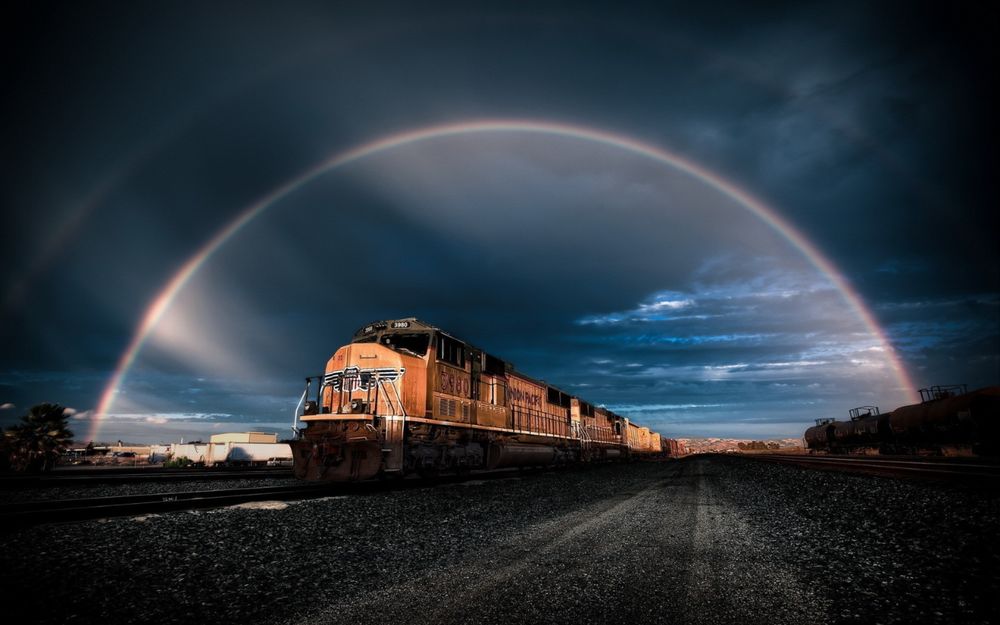 Обои для рабочего стола Тепловоз с грузовыми вагонами, идущий по железнодорожному полотну на фоне пасмурного синего вечернего неба и яркой дугообразной радуги