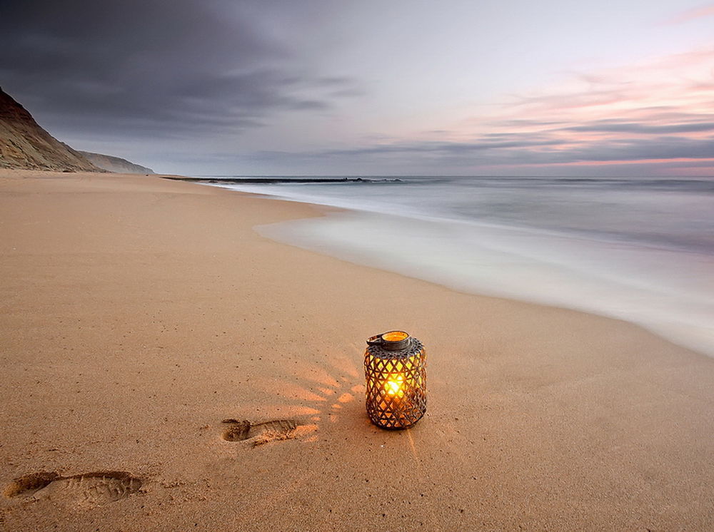 Обои для рабочего стола Декоративный фонарь в металлической сетке с горящей внутри свечой, стоящий на песчаном берегу моря рядом со следами человека, фотография ELOP