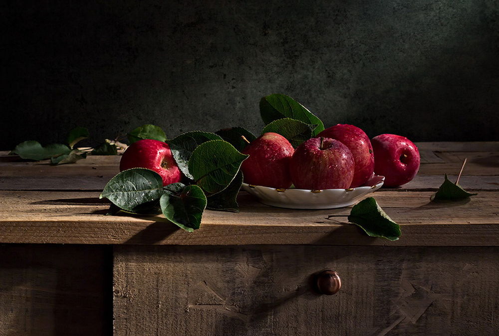 Обои для рабочего стола На деревянных неструганных досках стоит тарелка с красными, спелыми яблоками в капельках воды, рядом лежат темно-зеленые листочки, фотография Светланы Лебедевой