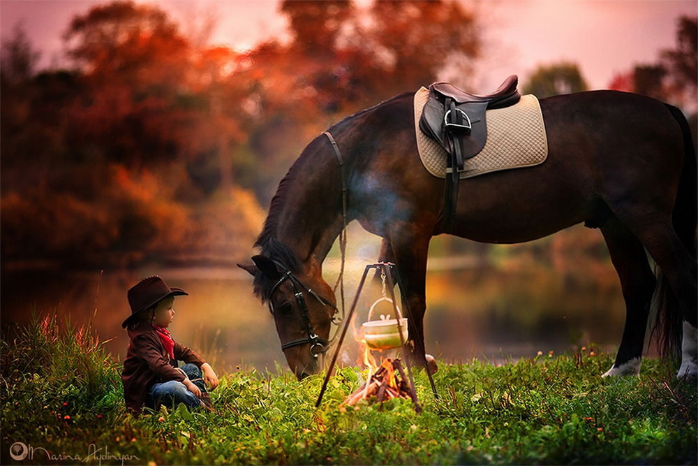 Обои для рабочего стола Мальчик в ковбойской шляпе и одежде, сидящий на зеленой поляне на берегу водоема, внимательно наблюдает за варящейся на костре ухой, рядом стоит статный гнедой конь, пощипывающий травку, фотография Марины Айдинян