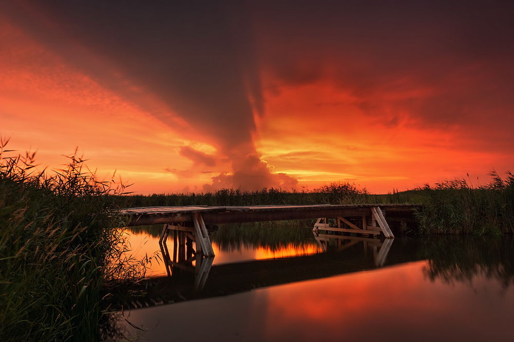 Обои для рабочего стола Деревянный мост, проложенный через неглубокую реку на фоне красивого желто-красного заката, отражающегося в зеркальной глади воды, фотография Adam Widowski