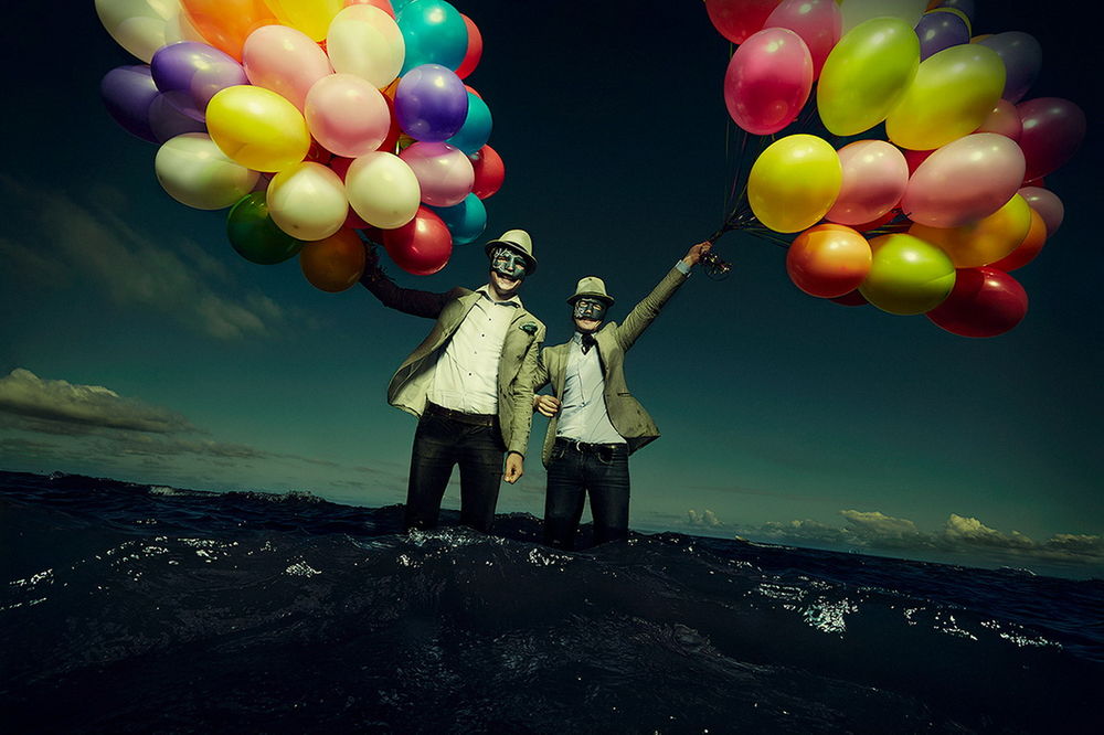 Обои для рабочего стола Двое мужчин в шляпах и масках, стоящие по колено в морской воде держат в руках большое количество разноцветных, надувных воздушных шариков, фотография Козин Ждет четвертого Всадника