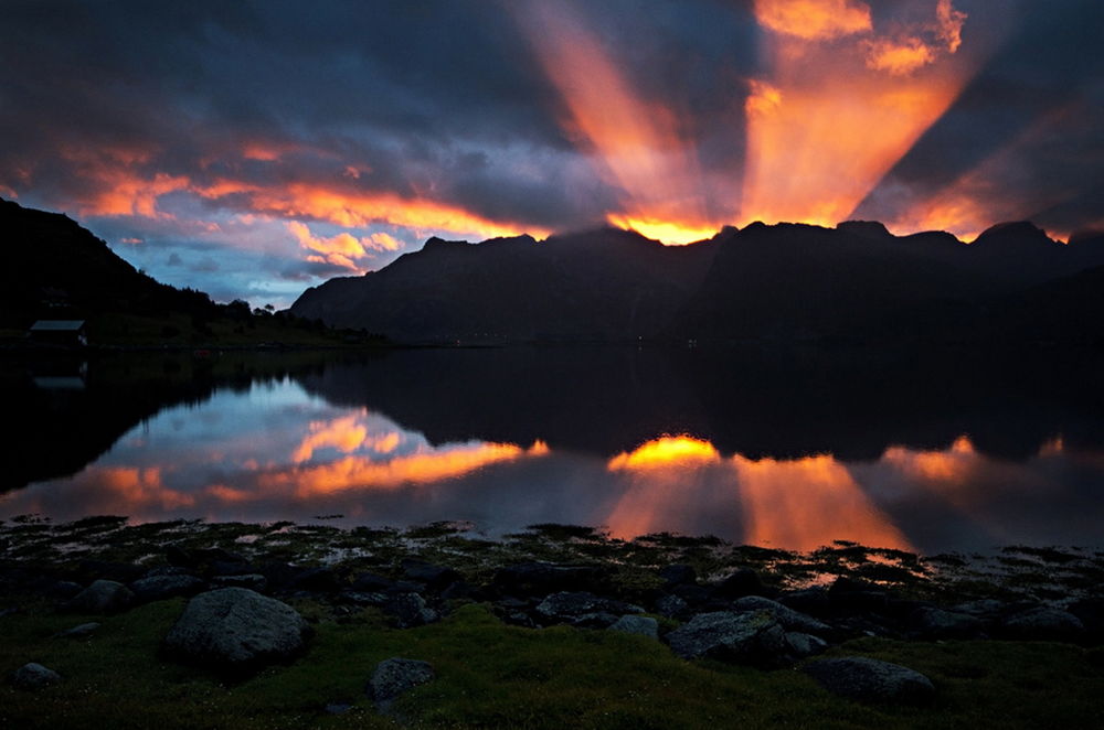 Обои для рабочего стола Лучи восходящего из-за гор утреннего солнца как прожектором осветили небо с темными облаками, отразившись в темной водной глади горного озера, фотография Andrey Bondarev