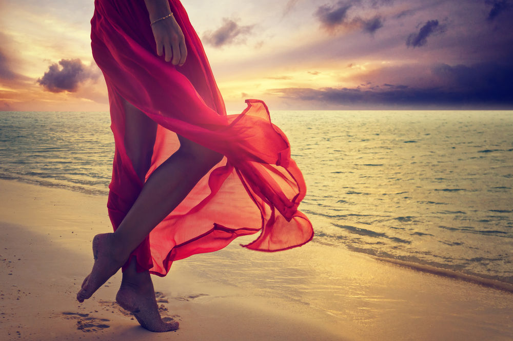 Обои для рабочего стола Девушка в красном платье босиком стоит на берегу моря