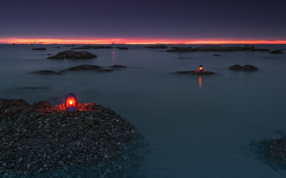 Обои для рабочего стола Горящие красным светом фонари, стоящие на валунах в прибрежной морской воде и расположенным на линии горизонта маяке на фоне полоски багряного заката и пасмурного неба