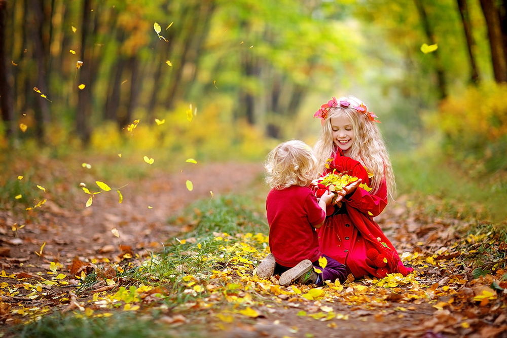 Обои для рабочего стола Белокурые брат и сестра, одетые в красную одежду, стоящие на коленях на лесной дорожке, усыпанной осенними листьями, держащие в руках, сплетенных вместе, горсточку таких же листьев, фотография Светланы Квашниной