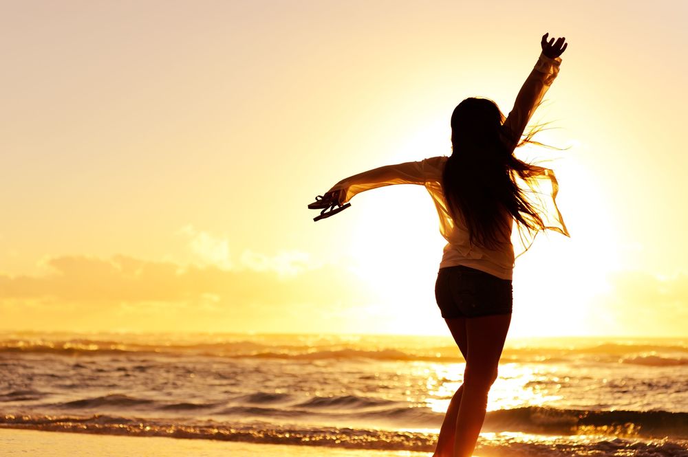 Обои для рабочего стола Девушка стоит на берегу моря, раскинув руки, на фоне заходящего солнца