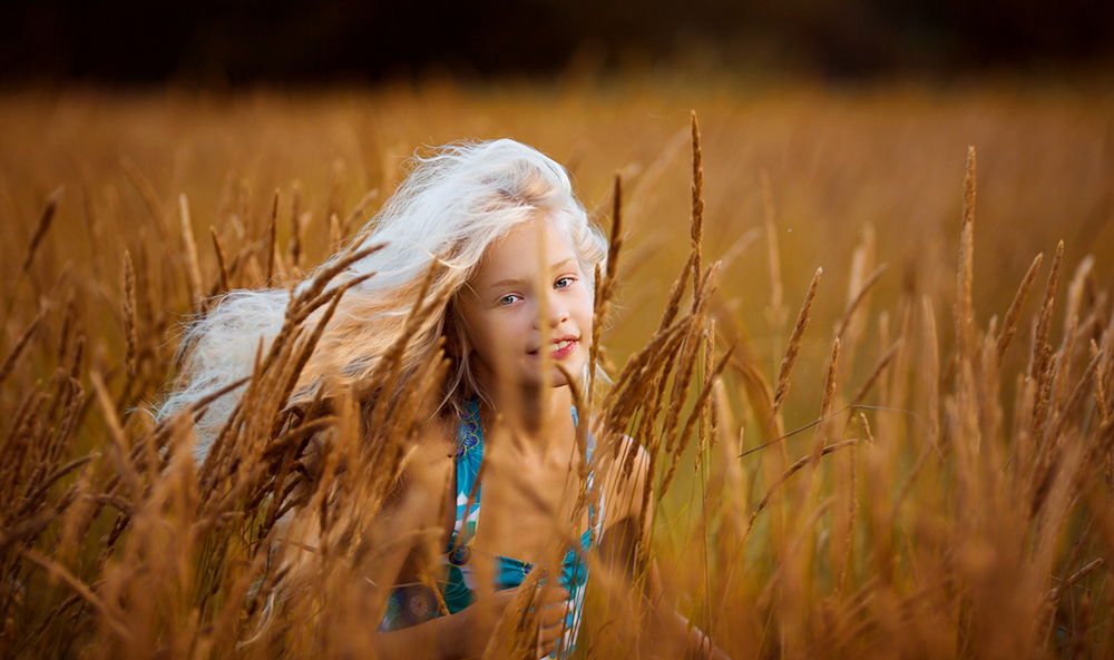 Обои для рабочего стола Милая, улыбающаяся девочка с длинными светлыми волосами, одетая в летний цветной сарафан, находящаяся в поле с колосьями созревшей пшеницы, фотография Светланы Квашниной