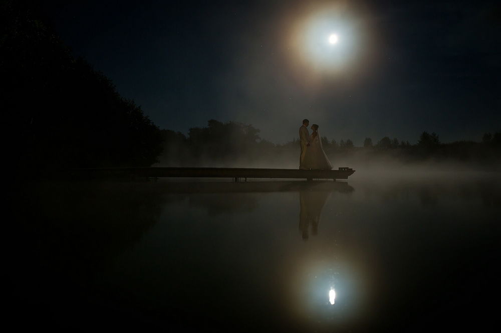 Обои для рабочего стола Романтическое свидание женщины в свадебном платье и мужчины в белом костюме, стоящих на деревянном мостике на озере, с поднимающейся над водной гладью легкой туманной дымкой на фоне ночного звездного неба и ярко светящейся луны, фотография Светланы Голицыной