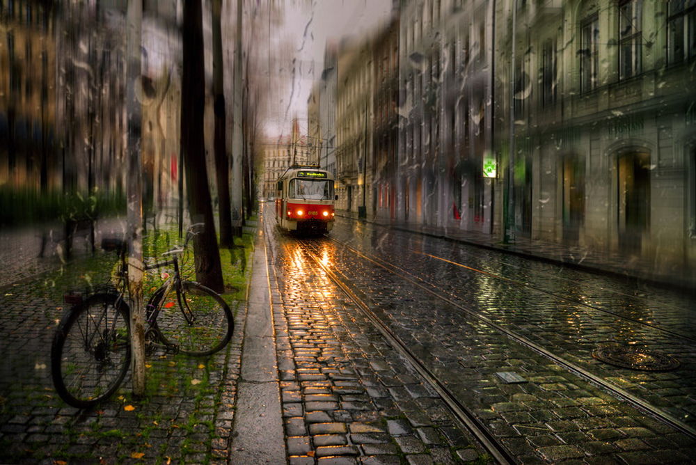 Обои для рабочего стола Трамвай с включенными фарами, идущий по вечерней, дождливой городской улице, к осветительной опоре, стоящей рядом с трамвайными путями прислонен велосипед, автор Эдуард Гордеев