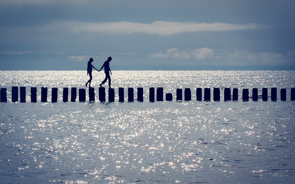 Обои для рабочего стола Парень и девушка, держась за руки идут по бетонным столбикам в прибрежной морской воде