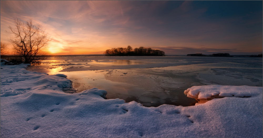 Обои для рабочего стола Заходящее за линию горизонта солнце осветило своими лучами снежный берег озера, поверхность которого покрылась тонким слоем льда