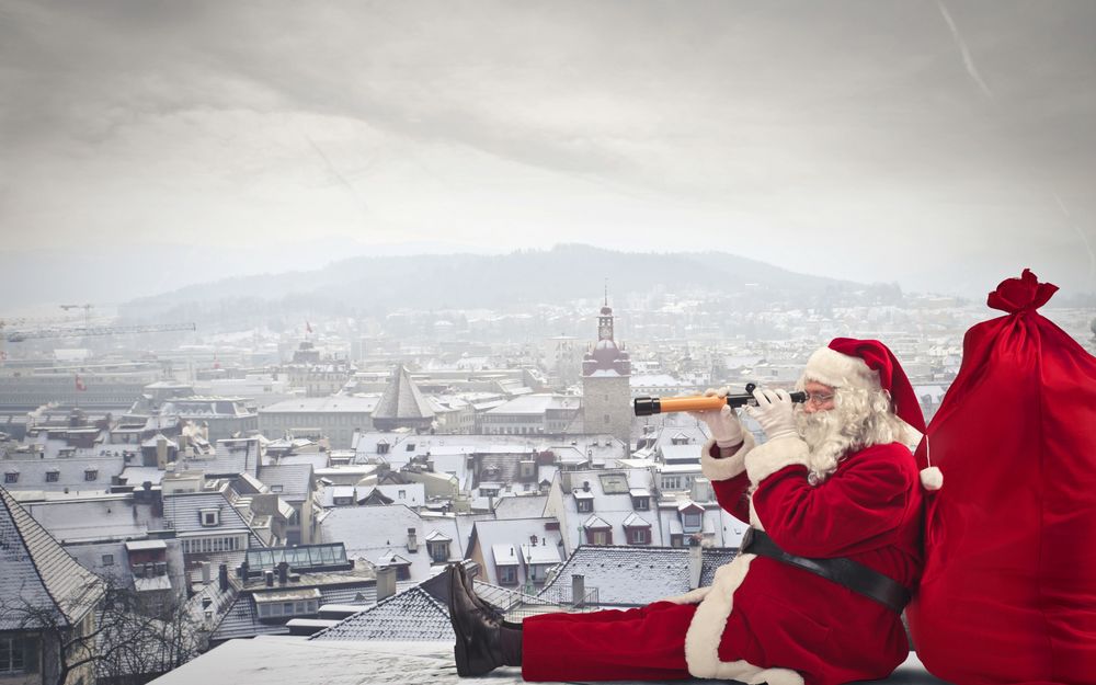 Обои для рабочего стола Санта Клаус сидит на крыше дома облокачиваясь на красный мешок и смотрит в подзорную трубу на фоне зимнего города