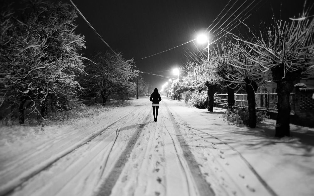 Обои для рабочего стола Высокая, стройная черноволосая девушка, идущая по дороге со свежевыпавшим снегом, по обочинам стоят деревья и уличные. ярко светящие фонари на фоне ночного неба
