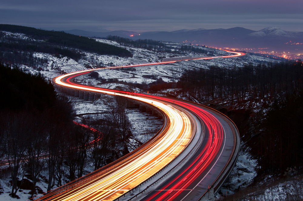 Обои для рабочего стола Светящиеся полосы от фар, проходивших на огромной скорости автомобилей на серпантине дороги, проложенной в горной местности на фоне вечернего, пасмурного неба