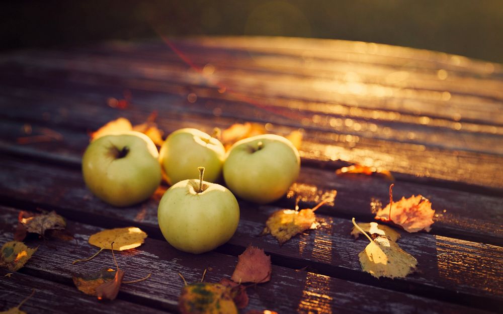 Обои для рабочего стола Четыре яблока лежат на деревянном столе, рядом разбросаны осенние желтые листья