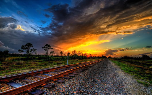 Обои на рабочий стол Заброшенное железнодорожное полотно с ржавыми рельсами, тянущееся к линии горизонта на фоне заката на небе с красивыми, разноцветными облаками