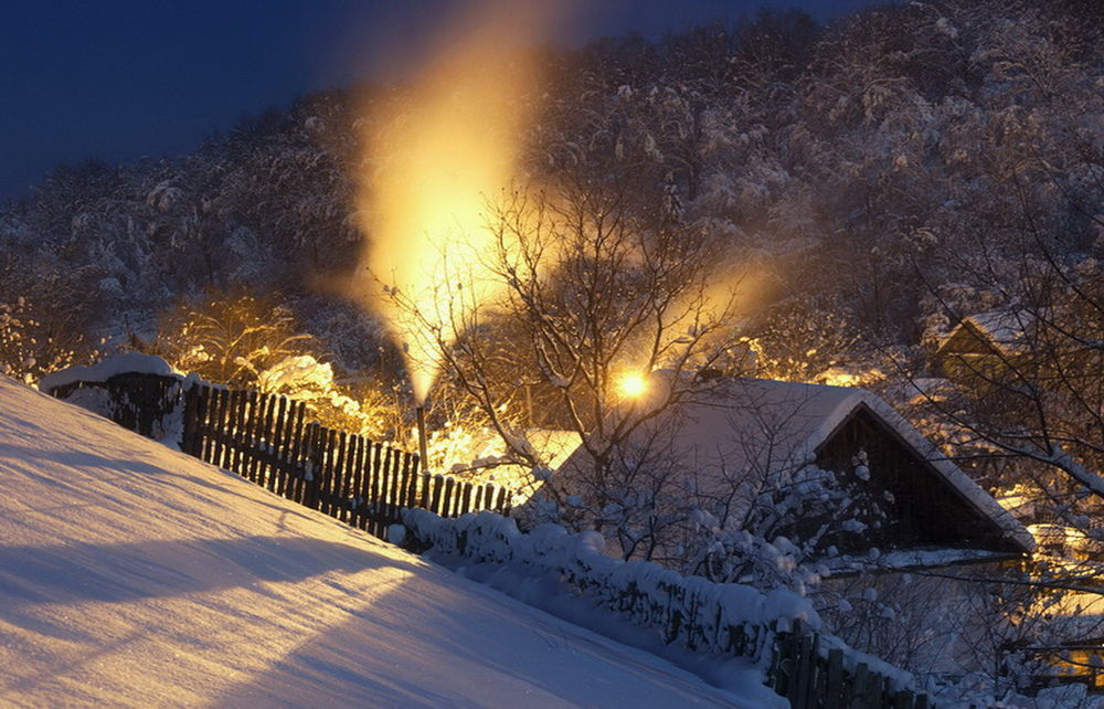 Обои для рабочего стола Небольшие деревянные домики, стоящие на окраине деревни, утопающие в снегу, освещенные электрическим освещением и ярким пламенем от горевшего газа, вырывающегося из металлической трубы