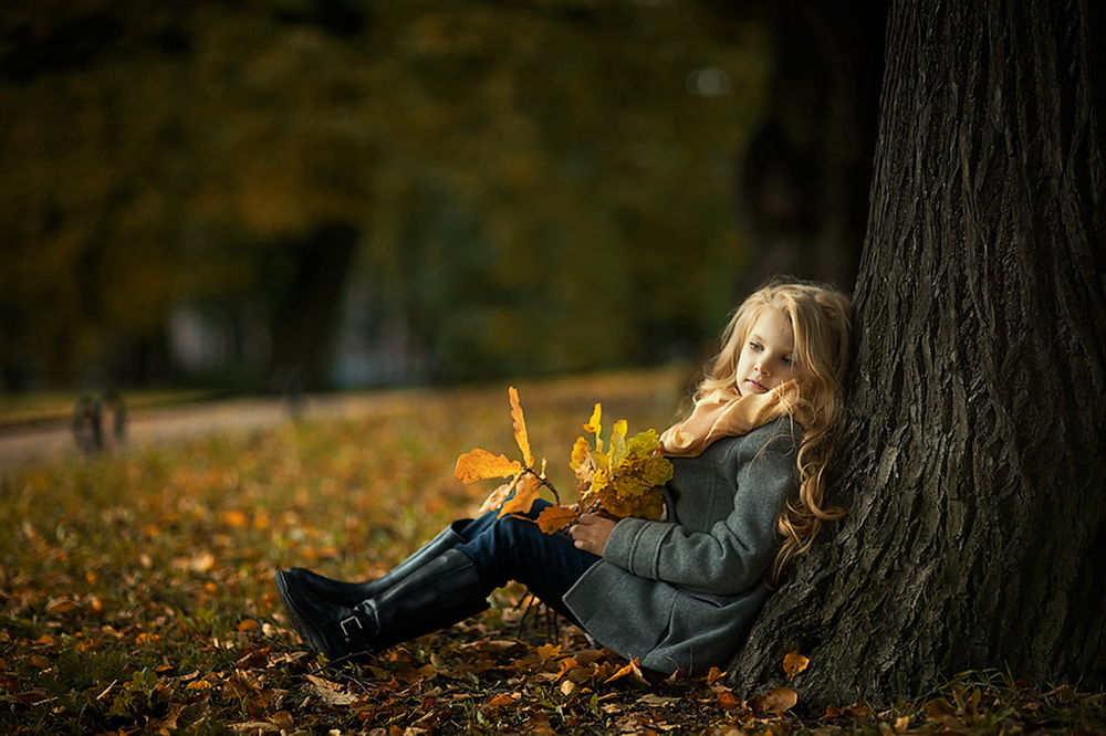 Обои для рабочего стола Милая, белокурая девочка с длинными волосами прислонилась спиной к дереву, сидя на осенних листьях и держа охапку таких же листьев в своих руках