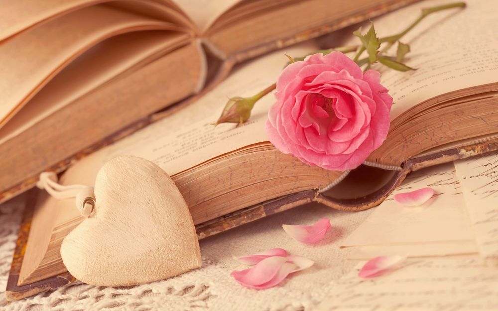 Обои для рабочего стола Кулон в виде сердца лежит на раскрытой книге, рядом розовая чайная роза с которой осыпаются лепестки