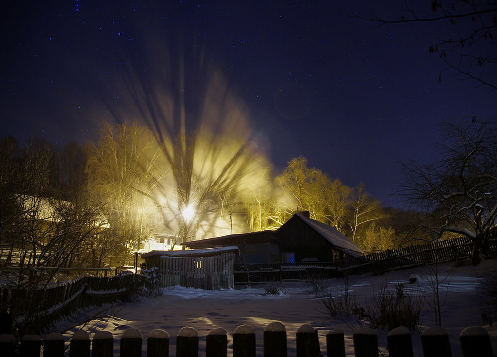 Обои для рабочего стола Горящее газовое пламя, вырывающееся из металлической трубы осветило деревья и деревянные дома, занесенные снегом на фоне ночного, звездного неба, фотограф Сергей Шляга