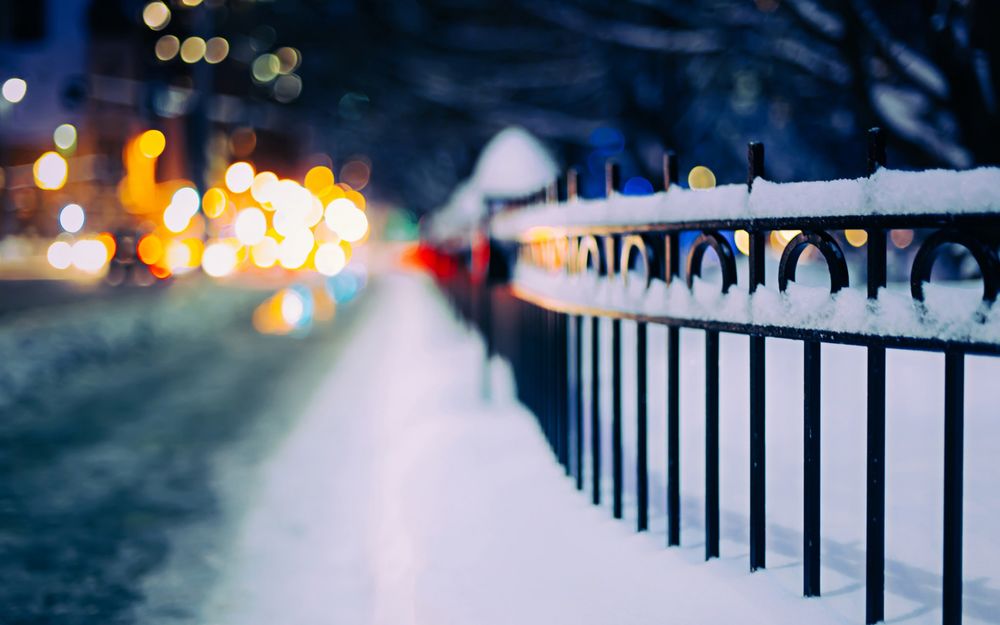 Обои для рабочего стола Дорога вдоль железной ограды, покрытой снегом, на фоне огней города