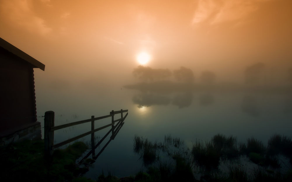 Обои для рабочего стола Деревянный сарай, стоящий на берегу озера на фоне утреннего восходящего солнца на небосклоне, затянутым густой, туманной пеленой