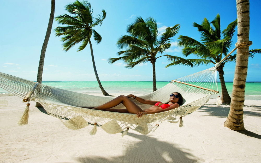 Обои для рабочего стола Девушка в красном купальнике отдыхает в гамаке на берегу красивого острова с рядом растущими пальмами