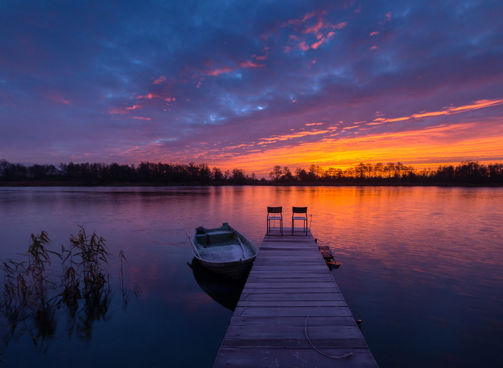 Обои для рабочего стола На деревянном причале, расположенном на озере, стоят два складных стула, рядом пришвартована лодка с лежащими в ней веслами на фоне заката на вечернем небосклоне с перистыми, разноцветными облаками, фотограф Mariuszbrcz