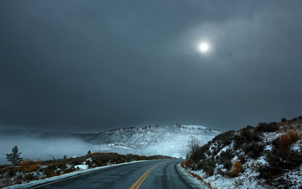 Обои для рабочего стола Асфальтовая дорога, проходящая по заснеженной холмистой местности на фоне ночного неба с взошедшей луной