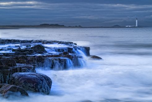 Обои на рабочий стол Камни на берегу моря, с которых течет вода, на линии горизонта маяк