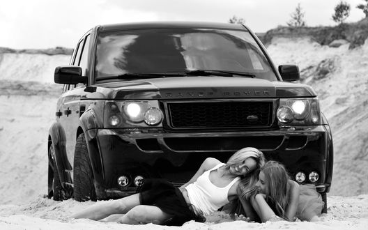Обои на рабочий стол Девушки лежат на песке у автомобиля Range Rover / Рэнж Ровер черного цвета и улыбаются