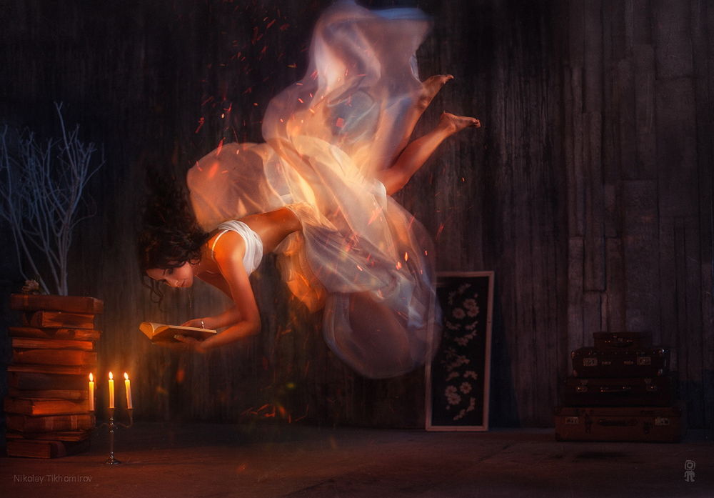 Обои для рабочего стола Темноволосая девушка в пышном, белом одеянии, парящая над полом в комнате, читающая книгу при горящих свечах, стоящих возле стопки книг, автор Николай Тихомиров