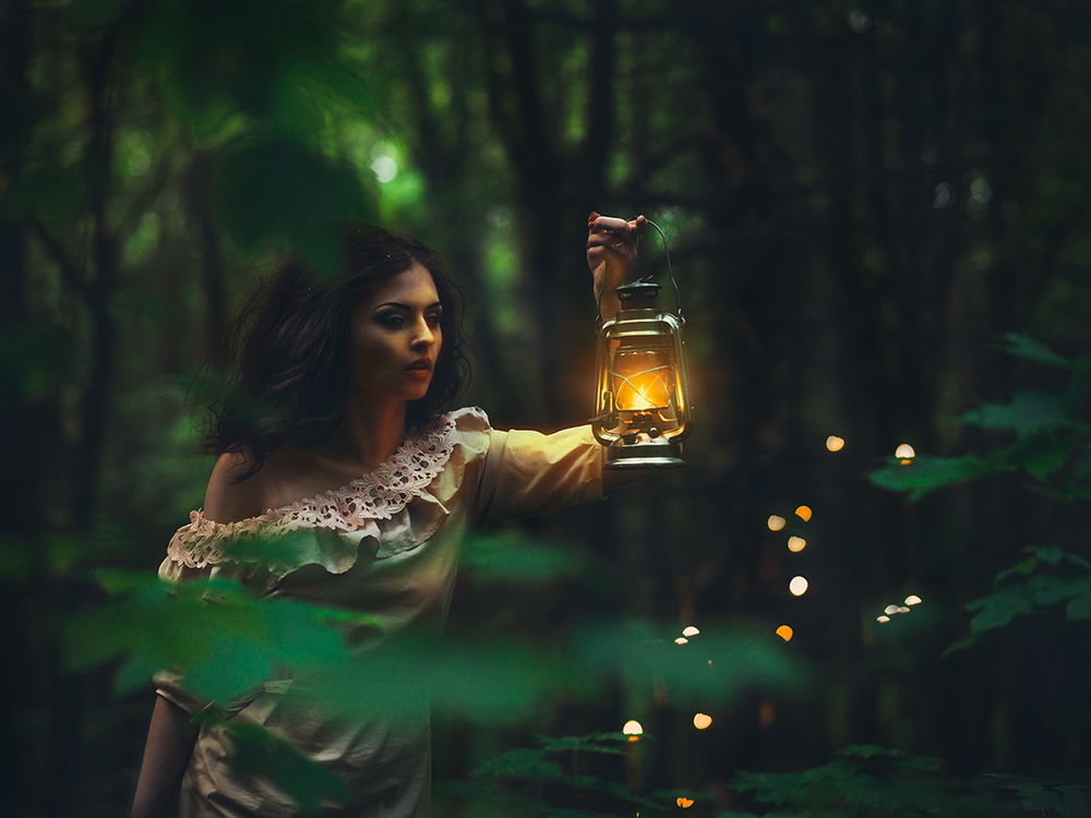 Обои для рабочего стола Красивая, темноволосая девушка, держащая в руке перед собой горящую, керосиновую лампу, идет по лесной чащобе в окружении бликов, автор Тарас Стельмах