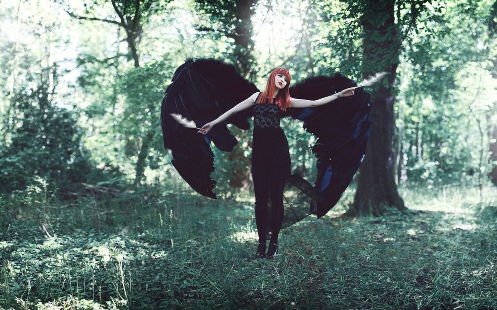 Обои для рабочего стола Девушка с черными крыльями и с двумя перьями в руках стоит на фоне деревьев