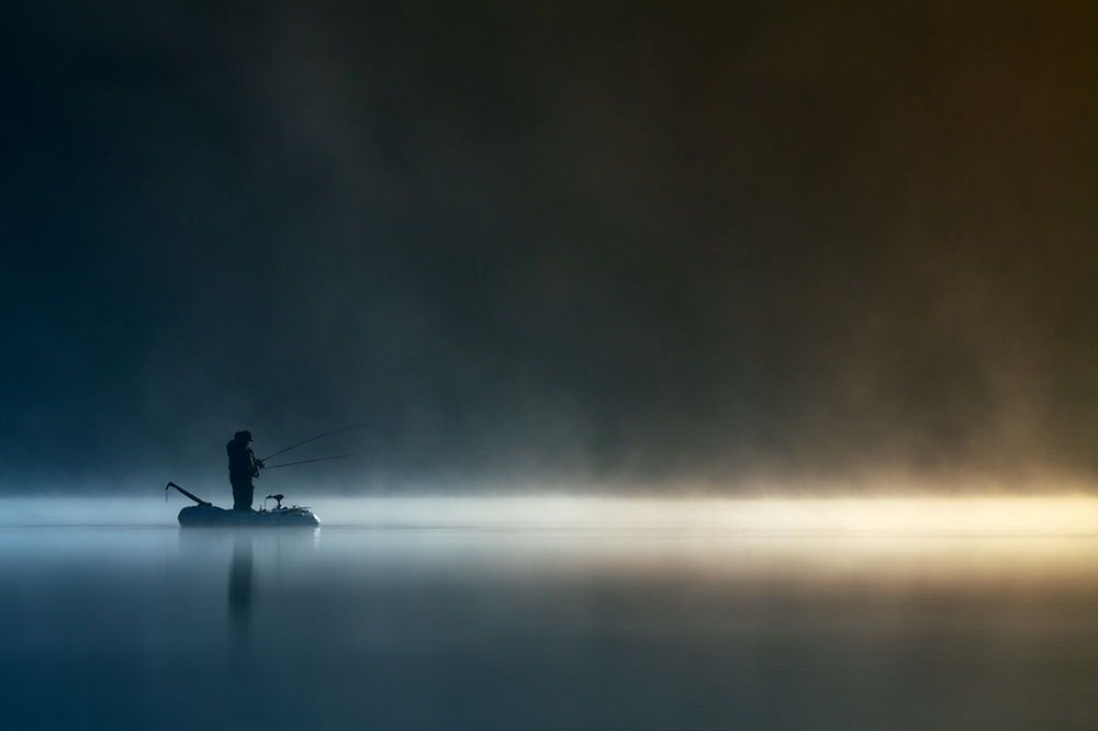 Обои для рабочего стола Рыбак, стоящий в надувной резиновой лодке, вышедший на утренний клев на реке, покрытой легким туманом