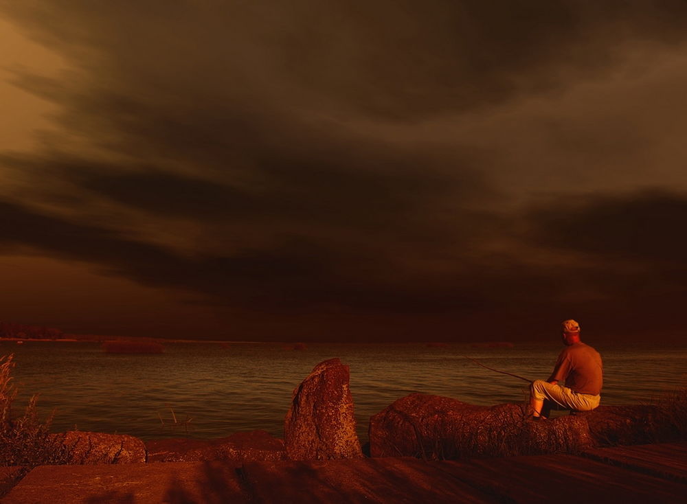 Обои для рабочего стола Рыбак, сидящий на камнях на берегу озера с удочкой в руках, освещенный лучами заходящего солнца на фоне темного, предгрозового неба