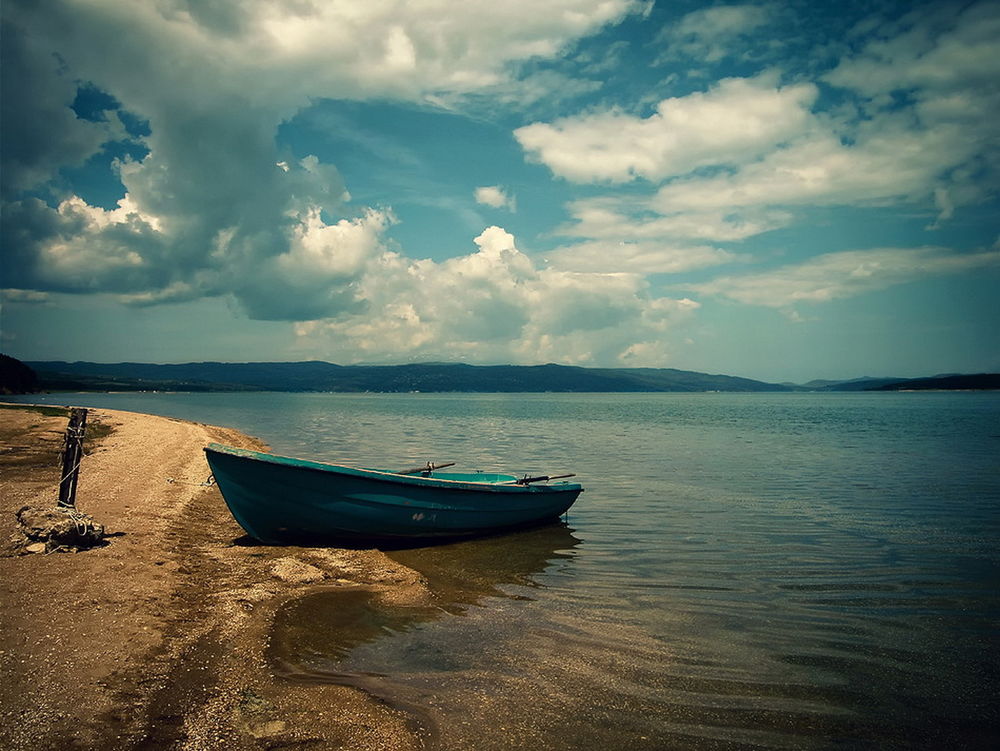 Обои для рабочего стола Лодка, стоящая у песчаного берега озера на фоне синего неба и белых кучевых облаков