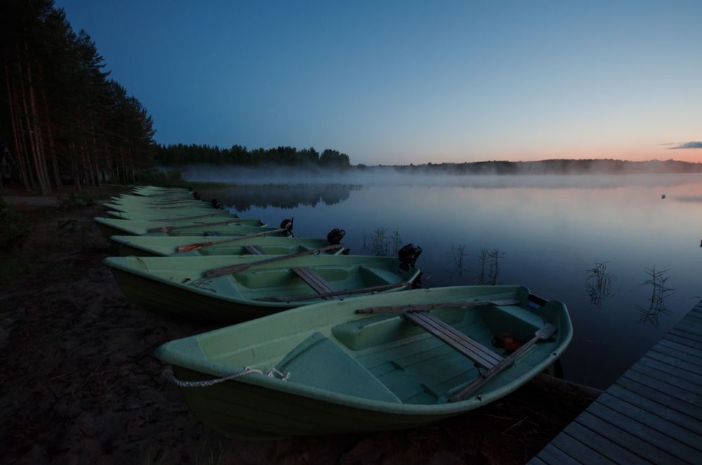 Обои для рабочего стола Лодки, стоящие на берегу озера невдалеке от деревянного причала на фоне утреннего, безоблачного небосклона с поднимающейся от поверхности воды туманной дымки