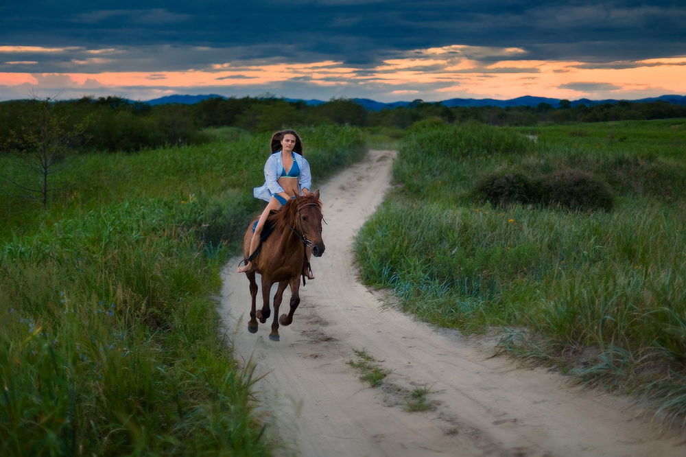 Обои для рабочего стола Девушка в легкой голубой рубашке, сидящая верхом на гнедой лошади, скачущей по песчаной дороге среди зеленого поля