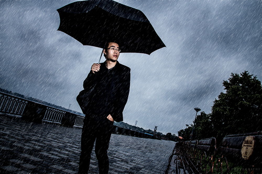 Обои для рабочего стола Мужчина азиатской внешности, держащий в руке черный зонтик, идущий под сильным дождем по брусчатой, городской мостовой