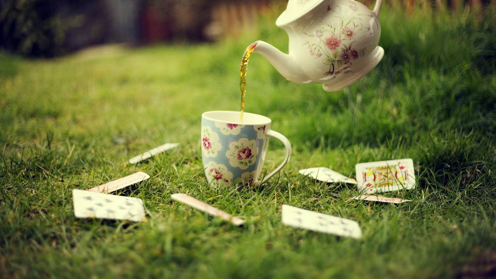 Обои для рабочего стола Из заварного чайника льется чай в кружку, стоящую на зеленой траве в окружении игральных карт