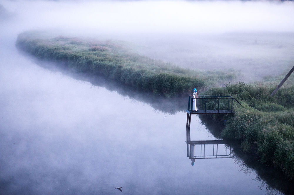 Обои для рабочего стола Босоногая девушка в длинном белом платье, стоящая на небольшом, деревянном мостике на берегу реки среди густых, зеленых кустов на рассвете с обволакивающим водную поверхность и небо туманом